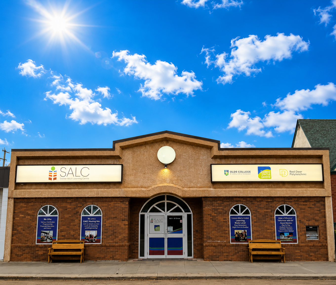 Front view of SALC learning centre—a brick building with large illuminated signs, arched windows, glass doors, and yellow benches outside under a blue sky.